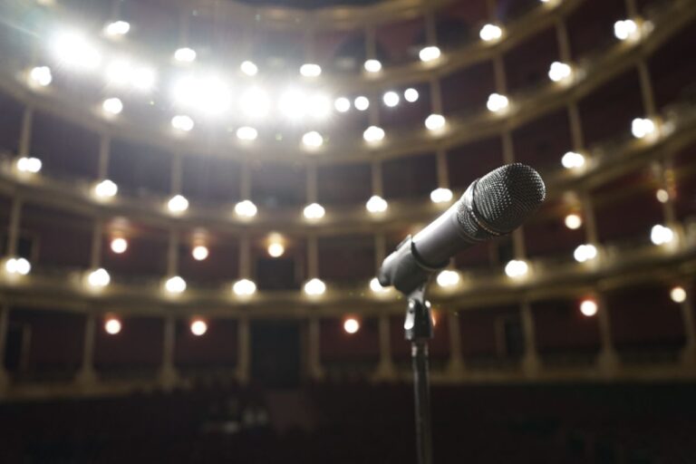 Close-up of a microphone on stage with bright theater lights in the background.